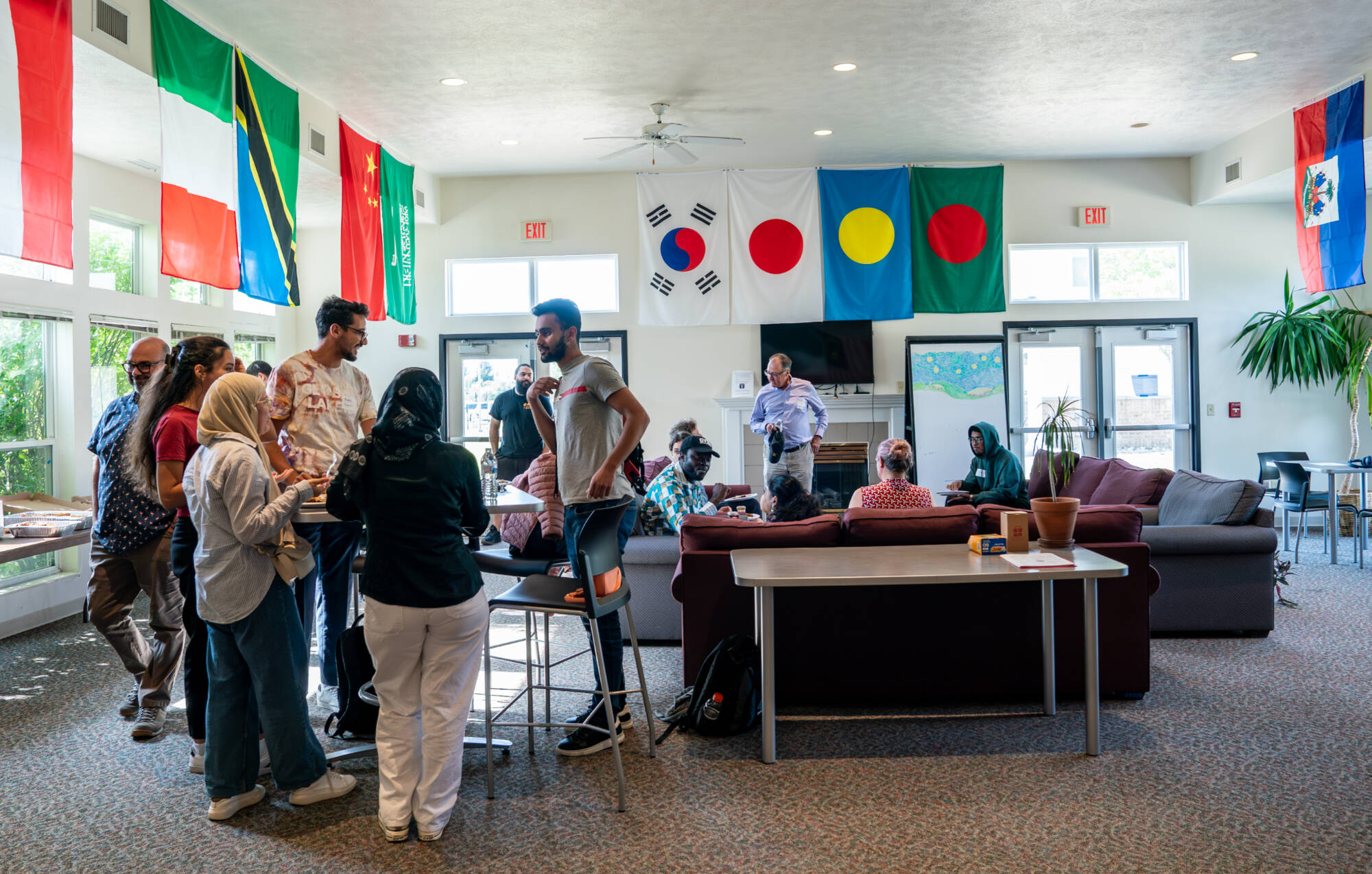Students, faculty, and staff gather for conversation during a College of Computing mixer at GVSU, with international flags displayed across the room and groups seated and standing together in a welcoming space.